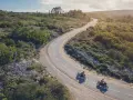 Due moto KTM sfrecciano su una strada tortuosa tra colline verdi. Un'avventura che celebra il KTM Connect.