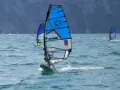 Osoba na desce windsurfingowej z niebieskim żaglem na wzburzonym morzu.