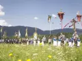 Menschen in Tracht und Uniformen nehmen an einer Prozession teil, die den Fronleichnam feiert. Fahnen und ein Kreuz werden getragen.