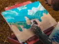 Un artiste tatoué est en train de peindre à la gouache sur toile un paysage montagneux sous un ciel bleu et nuageux.