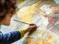 Une artiste restaure une fresque ancienne, sa main tenant un pinceau pour la préparation peinture. La scène montre une figure religieuse aux mains jointes.
