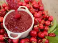 Cucharón vierte mermelada de cerezas casera en un cuenco blanco, rodeado de cerezas frescas y hojas verdes.