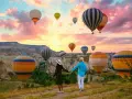 A couple holds hands, gazing at a sky filled with hot air balloons over the unique landscape of western Turkey at sunrise.