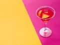 Elegancki cosmopolitan drink co zamiast żurawiny, z pomarańczą i lodem, na tle żółto-różowym.