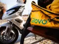 Żółta torba z logo Glovo na ławce obok białego skutera. Czeka na kolejnego kuriera Glovo.
