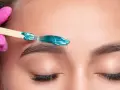A pink-gloved hand applies turquoise wax to an eyebrow. This close-up shows the contrast between hot wax vs cold wax application for brow shaping.