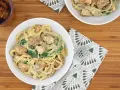 Delicioso plato de pasta alfredo con trozos de pollo y espinacas, servido en un tazón blanco sobre una tabla de madera.