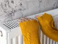Dwie dłonie w żółtych swetrach ogrzewają się przy białym grzejniku. Stalowy czy aluminiowy?