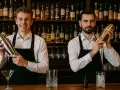 Dwóch barmanów w czarnych fartuchach i białych koszulach trzyma shakery do drinków na tle półek z butelkami alkoholu.