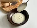 Una cucchiaiata di cremosa fonduta di parmigiano cade in una ciotola. Sul tagliere, un pezzo di Parmigiano Reggiano e un coltellino.