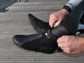 Mężczyzna zakłada czarne buty neoprenowe na drewnianym pomoście przy wodzie.