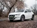 Biały Peugeot e-208 na brukowanej ulicy, z drzewami w tle.