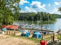 Mazury pod namiot: namioty i żaglówki przy pomoście, zielone drzewa i błękitne niebo z chmurami.