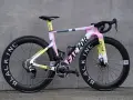 A pink, yellow, and gray Factor road bike with black wheels against a gray wall.