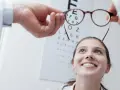 Okulista trzyma okulary przed twarzą uśmiechniętej kobiety na tle tablicy Snellena.