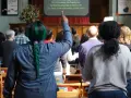 A woman with green hair raises her hand, perhaps inspired by the lyrics on screen, offering chant ideas for the congregation.