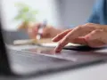 Dłoń na touchpadzie laptopa, w tle notatnik i długopis.