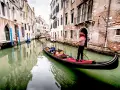 Gondoliere in abito a righe guida una gondola con turisti lungo un canale veneziano. Chissà quanto costa un giro in gondola a Venezia per vivere questa magia.