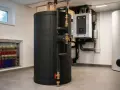 Duży, czarny bufor ciepła w kotłowni, obok białego zasobnika i kotła. Widać instalację hydrauliczną.