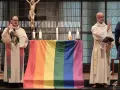 Zwei Geistliche stehen vor einem Altar mit einer Regenbogenfahne. Die Kirche feiert die gleichgeschlechtliche Ehe.