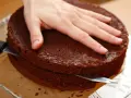 Eine Hand hält einen Schokoladenkuchen, während ein Messer ihn waagerecht durchschneidet, um den tortenboden zu teilen.