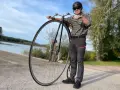 Man med hjälm och solglasögon står bredvid en cykel med stort framhjul vid en sjö.