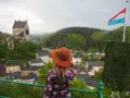 Mujer con sombrero marrón admira un castillo y un pueblo, con la bandera de Luxemburgo ondeando. ¡Un paisaje digno de un buen queso Muenster!