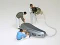 Miniaturowi pracownicy zajmują się naprawą aparatów słuchowych. Jeden z nich przykręca element, drugi czyści słuchawkę, a trzeci pracuje nad aparatem.