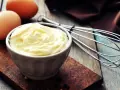 Un bol de mayonnaise onctueuse, des œufs et un fouet, prêts à faire une mayonnaise maison.