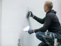 Mężczyzna w czarnej bluzie i rękawiczkach wykonuje tynkowanie ścian, używając kielni do rozprowadzania masy.