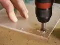 A drill bit is used to drill through acrylic. A hand guides the acrylic sheet as the drill creates a hole, showing it's possible to drill through acrylic.