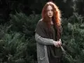 Rudowłosa dziewczyna w grubym swetrze i szalu, otoczona ciemną zielenią. Jej spojrzenie jest spokojne, a cała scena ma **low-key** klimat.