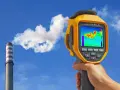 Ręka trzyma kamerę termowizyjną, która pokazuje ciepło wydobywające się z komina fabrycznego.
