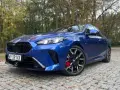 Błękitne, sportowe niemieckie auto BMW z czarnymi felgami i czerwonymi zaciskami hamulcowymi stoi na brukowanej drodze wśród jesiennych drzew.