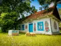 Malowany dom w Zalipiu, z niebieskimi oknami i drzwiami, ozdobiony kwiatami. Obok studnia i snop siana. Cennik wizyty w tym malowniczym miejscu jest dostępny dla zwiedzających.