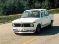Białe BMW E20 z kolorowymi pasami i napisem "turbo" na błotniku, z kierowcą za kierownicą, jedzie po asfaltowej drodze.
