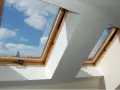 Dwa drewniane okna dachowe z obróbką, przez które widać błękitne niebo z chmurami.