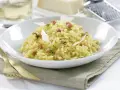 Un cremoso arroz a la milanesa, adornado con láminas de queso parmesano y hierbas frescas, servido en un plato blanco.