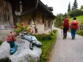 Due escursioniste affrontano il cammino San Vili, con zaini rossi e bastoncini. La strada, tra case rurali e verde, suggerisce una certa difficoltà.
