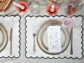 A table setting with a gold-colored tablecloth, white placemats with black scalloped edges, plates, cutlery, glasses, and a menu with illustrations.