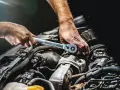 Mechanik dokręca śrubę kluczem w silniku samochodu. Widać jego dłonie i fragmenty silnika, być może podczas rutynowej kontroli lub naprawy.