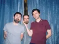 Trois hommes posent devant un rideau bleu. L'un d'eux, au centre, est un groupe musique espagnol année 2000.