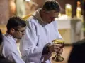 Szafarz, w białej albie, trzyma złoty kielich z hostiami. Obok niego młodszy mężczyzna w okularach.