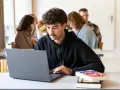 Ein junger Mann mit Schnurrbart konzentriert sich auf seinen Laptop, während er ein evangelisches Fernstudium Theologie absolviert.