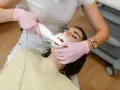 Dentystka w różowych rękawiczkach wykonuje zabieg piaskowania zębów. Czy piaskowanie zębów boli? Pacjentka ma otwarte usta i rozwierak.