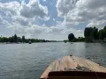 Blick vom Bug eines Holzbootes auf einen Fluss mit vielen Booten und grünen Ufern. Ein schöner Tag, fast wie ein Heidelberg See.