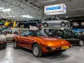 Pomarańczowa Mazda RX-7 błyszczy w muzeum samochodów, obok innych klasyków marki.