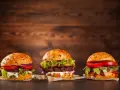 Trzy apetyczne burgery z różnymi dodatkami na drewnianym tle.