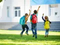 Drei Kinder mit Rucksäcken springen fröhlich vor einem Gebäude.