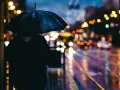 Person sous un parapluie noir, attendant sur un trottoir mouillé. Les lumières de la ville se reflètent sur la chaussée, créant de belles photos de pluie.